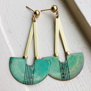 May include: A pair of gold and teal green earrings with a geometric design. The earrings are made of metal and have a semi-circular shape with a black line pattern.