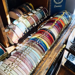 May include: A display of colourful leather bracelets with metal accents. The bracelets are arranged in rows on a wooden surface. The bracelets are all different colours, including brown, red, blue, green, and yellow. Some of the bracelets have metal clasps, while others have metal beads or studs.