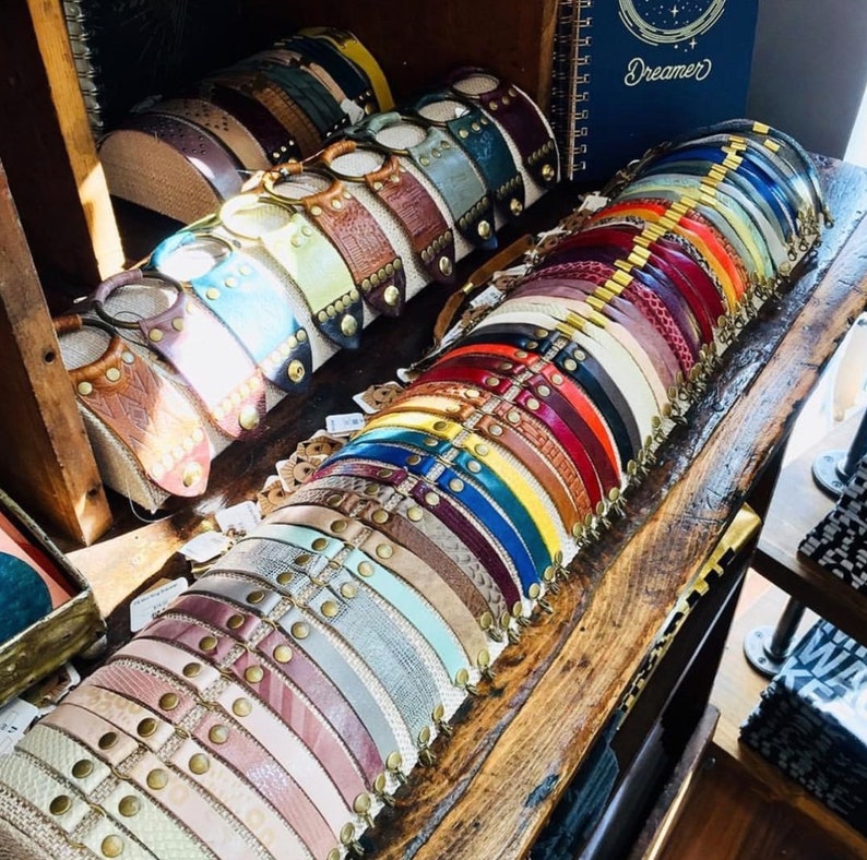 May include: A display of colourful leather bracelets with metal accents. The bracelets are arranged in rows on a wooden surface. The bracelets are all different colours, including brown, red, blue, green, and yellow. Some of the bracelets have metal clasps, while others have metal beads or studs.