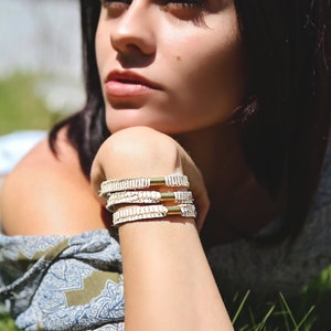 May include: A woman wearing a white and blue patterned shirt with three white leather bracelets with gold accents on her wrist.