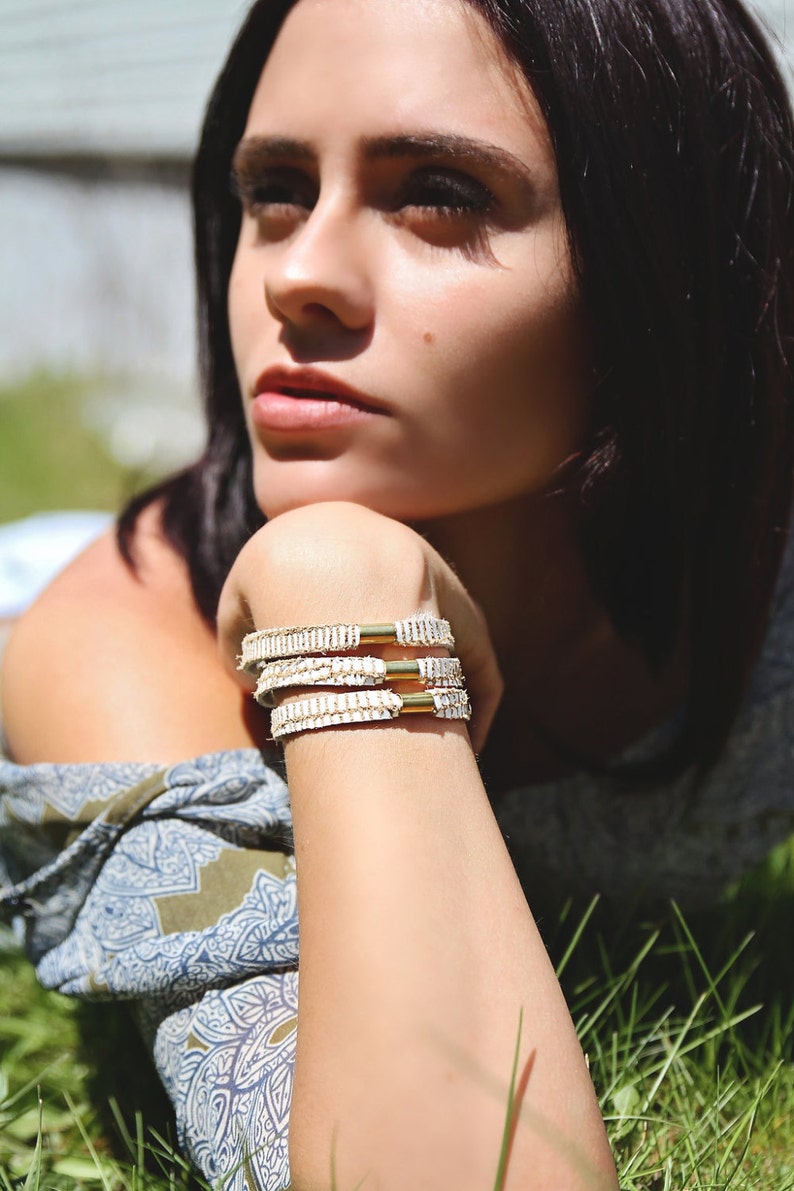 May include: A woman wearing a white and blue patterned shirt with three white leather bracelets with gold accents on her wrist.