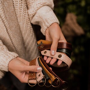 May include: A collection of leather keychains in various colors, including brown, tan, green, and yellow. The keychains are made of braided leather and have a metal ring at the end. The keychains are held in a person's hand.