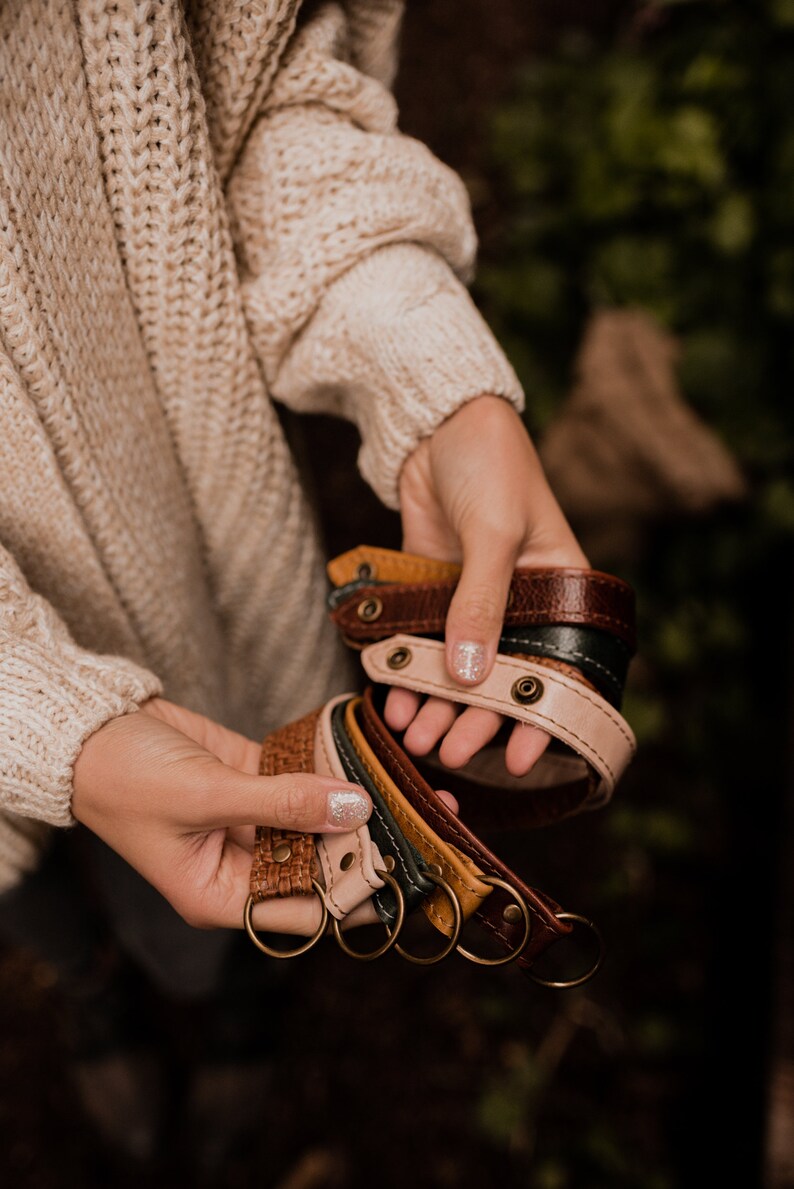 May include: A collection of leather keychains in various colors, including brown, tan, green, and yellow. The keychains are made of braided leather and have a metal ring at the end. The keychains are held in a person's hand.