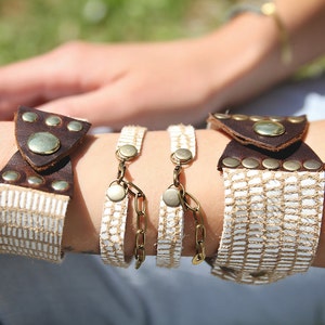 May include: A set of three bracelets with white woven fabric and brown leather accents. The bracelets feature decorative metal accents and closures.