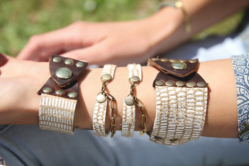 May include: A set of three bracelets with white woven fabric and brown leather accents. The bracelets feature decorative metal accents and closures.
