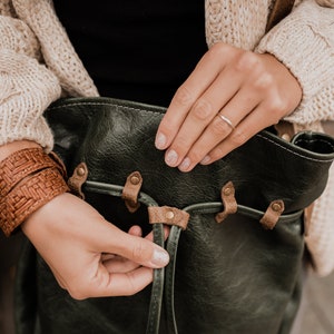 May include: A woman adjusts the strap of a dark green leather bag with brown leather accents. The bag has a woven brown leather strap.