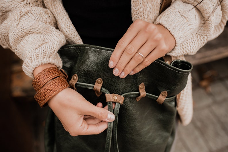 May include: A woman adjusts the strap of a dark green leather bag with brown leather accents. The bag has a woven brown leather strap.