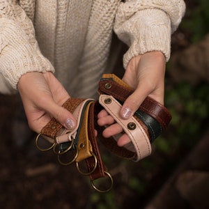 May include: A set of five leather keychains in various colors, including brown, tan, pink, and green. Each keychain has a metal ring and a decorative metal button.