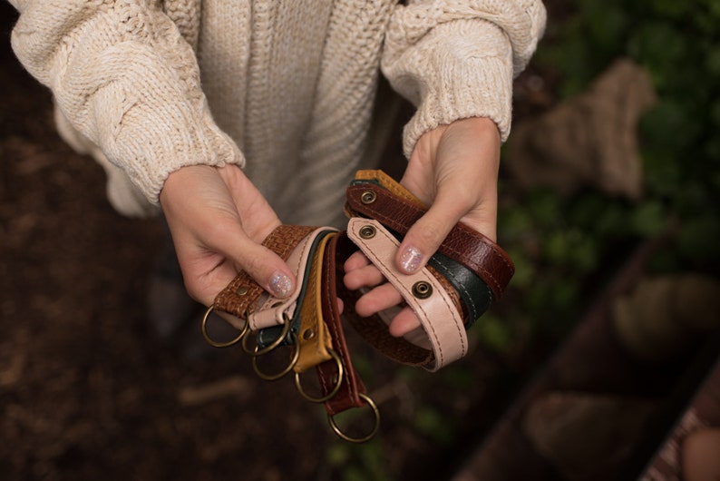 May include: A set of five leather keychains in various colors, including brown, tan, pink, and green. Each keychain has a metal ring and a decorative metal button.