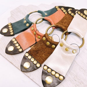 May include: Four leather keychains with gold rings and decorative brass studs. Each keychain has a different color of leather: teal, peach, brown, and white.