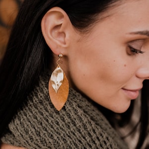 May include: A woman wearing a brown leather and gold leaf dangle earrings. The earrings are in the shape of a leaf and have a gold metal backing.