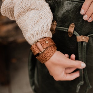 May include: A brown leather bracelet with a woven design and a metal ring. The bracelet is worn on a person's wrist.