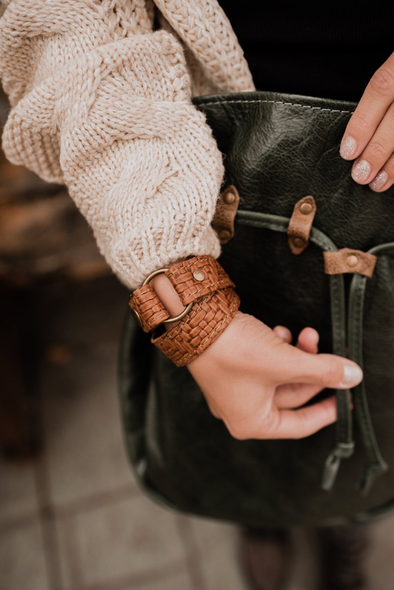 May include: A brown leather bracelet with a woven design and a metal ring. The bracelet is worn on a person's wrist.
