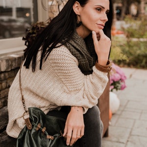 May include: A woman wearing a cream-colored knitted sweater, a green scarf, and black jeans sits on a wooden bench. She is holding a dark green leather bag with brown straps and buckles.