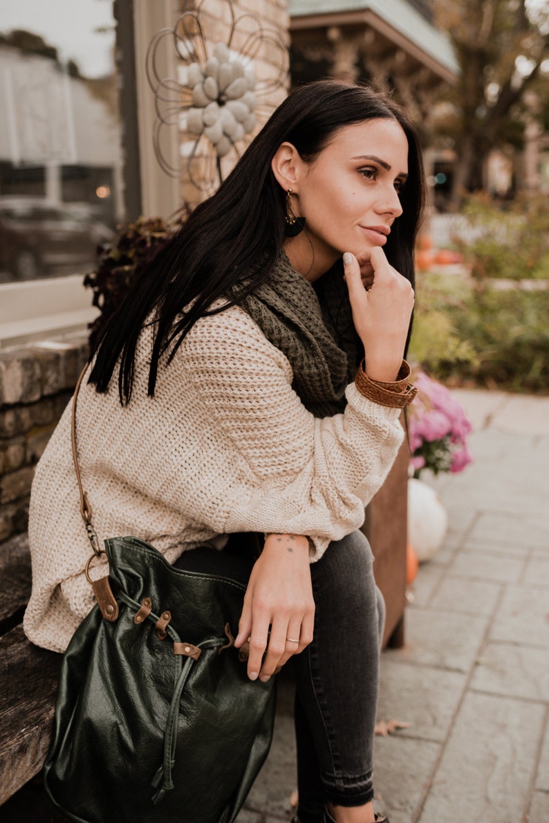 May include: A woman wearing a cream-colored knitted sweater, a green scarf, and black jeans sits on a wooden bench. She is holding a dark green leather bag with brown straps and buckles.