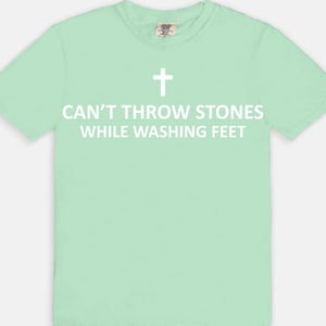 May include: A light green t-shirt with a white cross and the text "CAN'T THROW STONES WHILE WASHING FEET". The shirt has short sleeves and a crew neck.