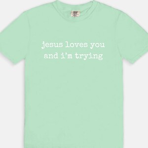 May include: A light green t-shirt with the text "jesus loves you and I'm trying" in white. The shirt has a crew neck and short sleeves. The fabric appears to be soft and comfortable.