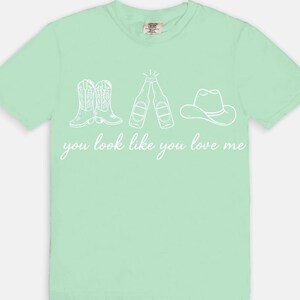May include: A light green t-shirt with a white graphic of cowboy boots, beer bottles, and a cowboy hat. The text "you look like you love me" is written below the graphic in a cursive font.