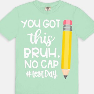 May include: A light green t-shirt with the text "YOU GOT this BRUH. NO CAP #testDay" in white. A yellow pencil graphic with a pink eraser and gray accents is on the right side of the shirt. The shirt is a comfortable, casual style.