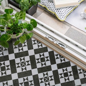 May include: A white tiled floor with a black and white geometric pattern. A white door with a silver handle is open, revealing a patio with plants, a book, and a pillow.