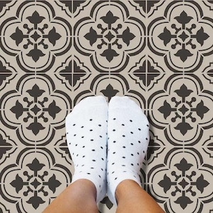 May include: A stencil featuring a repeating pattern of black floral designs on a beige background. The stencil is being used to create a decorative floor design. A pair of white socks with black polka dots are visible in the foreground.