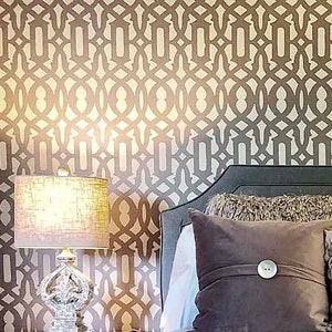 May include: A bedroom with a bed with a brown headboard and a white duvet. The bed has a brown throw blanket and several pillows. There is a small plant in a gray pot on a silver tray on the bed. The wall behind the bed is covered in a brown and white geometric wallpaper.