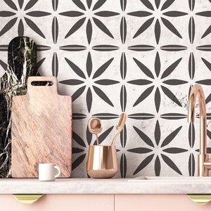 May include: A kitchen countertop with a white tiled backsplash featuring a geometric pattern of black flower shapes. The countertop has a white marble surface with a faucet, two cutting boards, a cup, and a copper utensil holder.