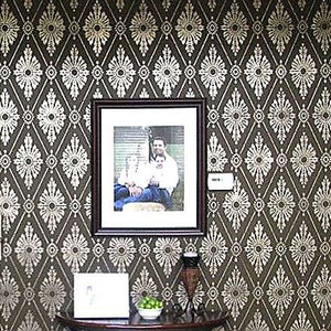 May include: A brown wall with a white geometric pattern. A framed photo of a family of three is hanging on the wall. A small table with a vase and a bowl of limes is in front of the wall.