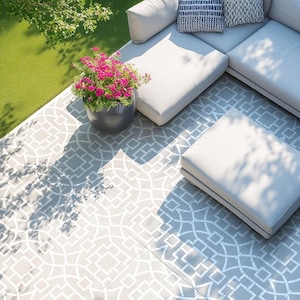 May include: An outdoor patio scene featuring a grey geometric patterned tile floor. A grey sectional sofa with white cushions and patterned pillows is visible. A dark grey pot with pink flowers sits nearby, with green grass in the background.