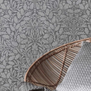 May include: Grey wallpaper with a repeating floral pattern of leaves and flowers. A woven wicker chair and a grey textured cushion are in the foreground. The wallpaper has a vintage, botanical design.