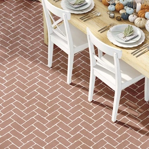 May include: A wooden dining table with two white chairs. The table is set with a white tablecloth, plates, silverware, and a centerpiece of fall gourds and candles. The floor is a herringbone pattern of brown tiles.