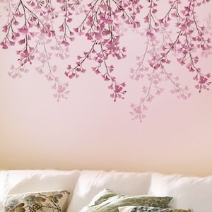 May include: A pink wall with a stencil of pink flowers hanging from branches. The stencil is in the upper portion of the image. The lower portion of the image shows a white couch with pillows.