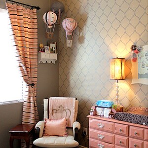 May include: A nursery room with a pink and white dresser, a rocking chair with a pink pillow, and a patterned wallpaper with a diamond design. There are hot air balloon decorations on the wall.