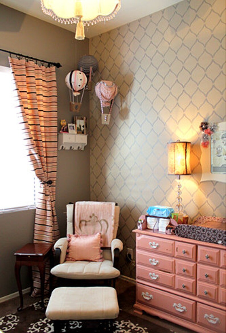 May include: A nursery room with a pink and white dresser, a rocking chair with a pink pillow, and a patterned wallpaper with a diamond design. There are hot air balloon decorations on the wall.