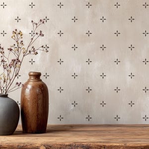 May include: A still life composition featuring two ceramic vases, one gray and one brown, with dried flowers. The background is a beige wall with a repeating dark brown cross pattern. The vases sit on a wooden surface.