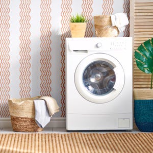 May include: A white washing machine is in a laundry room with a woven rug and baskets. The wall has a white background with vertical wavy lines in a reddish-brown color. A potted plant and folded towels are on top of the machine.