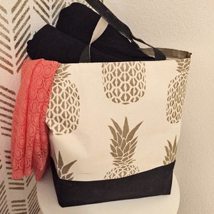 May include: A canvas tote bag with a cream background and large brown pineapple print. The bottom of the bag is black. Inside the bag are rolled black items and a coral-colored lace scarf. Black handles.