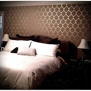 May include: A white bed with a brown headboard and white pillows. The wall behind the bed is painted a light brown color and has a gold stencil pattern.