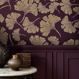 May include: Deep plum wallpaper featuring a repeating pattern of gold ginkgo leaves. In the foreground, a round white marble table holds a dark textured vase and a small stacked wooden box against a matching plum-coloured panelled wall.