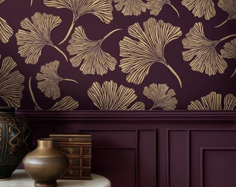 May include: Deep plum wallpaper featuring a repeating pattern of gold ginkgo leaves. In the foreground, a round white marble table holds a dark textured vase and a small stacked wooden box against a matching plum-coloured panelled wall.