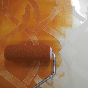 May include: A close-up of a paint roller with a white handle, being used to apply orange paint to a stencil on a wall. The stencil creates a repeating pattern of interlocking shapes.