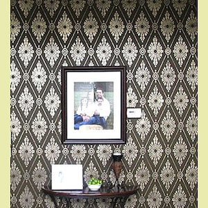 May include: A brown wall with a white diamond pattern stencil design. A framed photo of a family of three is hanging on the wall. A small table with a vase and a bowl of green limes is in front of the wall.
