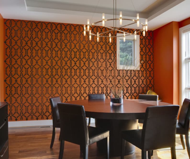 May include: A dining room with a round wooden table and six black leather chairs. The wall is painted orange and features a black geometric stencil pattern. A modern chandelier hangs above the table.