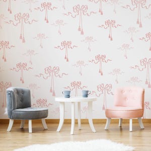 May include: Two small chairs, one grey and one pink, sit in front of a white table with two white cups. The wall behind the furniture is white with a repeating pattern of pink bows with tassels.