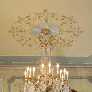 May include: A gold chandelier with crystal accents hangs from a ceiling with a decorative painted design. The design features a circular pattern of gold and green floral motifs.
