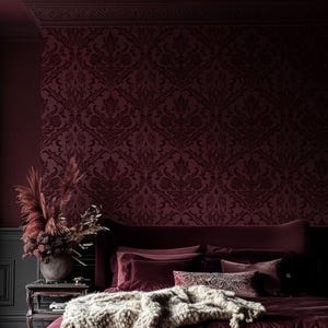 May include: A luxurious bedroom with a burgundy color scheme. The focal point is a bed with a velvet headboard, adorned with plush pillows and a textured throw. The walls feature a patterned wallpaper, and a vase of dried flowers sits on a side table.