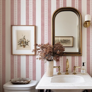 May include: A bathroom with a white marble countertop, gold fixtures, and a large arched mirror. The walls are decorated with a red and white striped wallpaper. A framed botanical print hangs on the wall, adding a touch of nature.