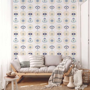 May include: A white wall with a repeating pattern of blue and gold eye designs. A wooden futon with pillows and a blanket is in front of the wall. A small wooden stool with decorative items and a woven basket are in the foreground.