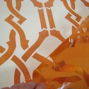 May include: A close-up of an orange stencil being peeled off a white surface. The stencil has a geometric pattern of interlocking shapes.
