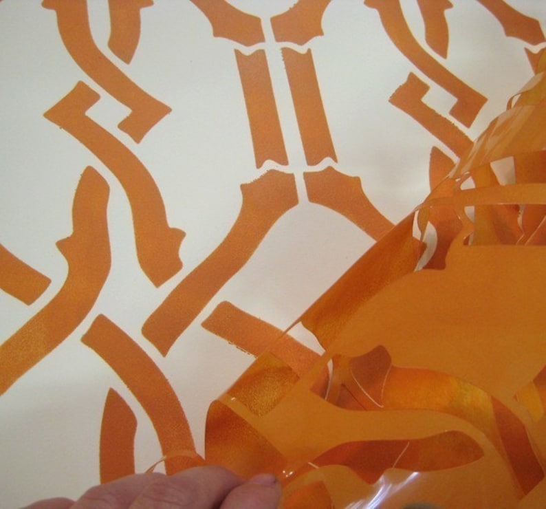 May include: A close-up of an orange stencil being peeled off a white surface. The stencil has a geometric pattern of interlocking shapes.